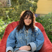 Shoshana, smiling, while reclining in a red chair in front of trees. Student-supplied profile photo due to COVID-19. Thank you, Shoshana!