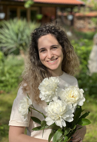Yoana smiling outside, wearing a white top and holding large white flowers.