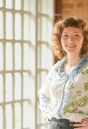 Jade smiling at the camera indoors, near a large window. She is wearing a floral button-up shirt.