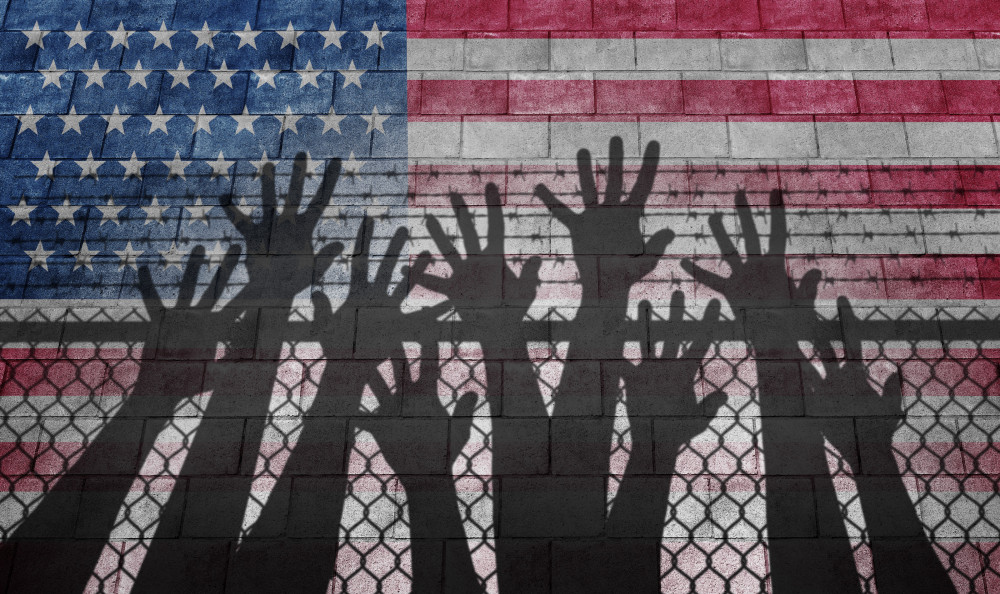 Shadow of hands reaching up over a chain link fence with an American flag as the background.