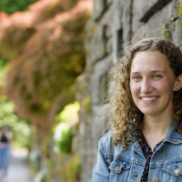 Mara, wearing a floral print shirt and denim jacket, smiling in front of a stone wall near the campus reflecting pool.