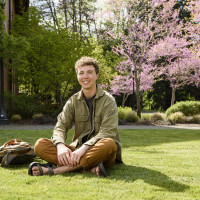 Max, sitting on the grass next to his backpack, smiling on a sunny day while wearing a green coll...