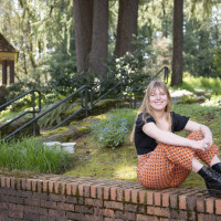 Jemma smiling outside, surrounded by trees, wearing a black top, orange patterned pants, and black boots.