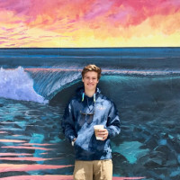 Max, holding a cup of coffee, standing in front of a wall with a painted mural of the ocean. Student-supplied profile photo due to COVID-...