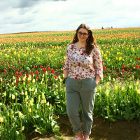 Sophie posing in front of a field of colorful wildflowers. She is wearing glasses, a shirt with flowers on it, and green pants.