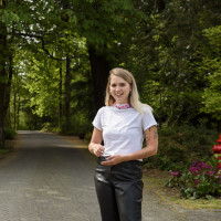 Elana, in black pants and a white t-shirt, smiling while standing in the tree-lined pathway betwe...