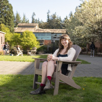 Bryn sitting in a chair outside with Watzek Library in the background. She is wearing a black dress with a white cardigan and brown boots.