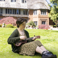 Mack sitting outside Manor House playing a ukulele. They are wearing a green floral dress with a dark green cardigan and black loafers.