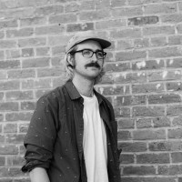 Black and white photo of Jonah. He is wearing a baseball hat, glasses, and a dark button up shirt open over a white tee.