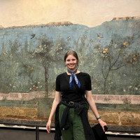 Claire Lyman posing in front of a mural wearing a black top, green pants, and a blue scarf around her neck.