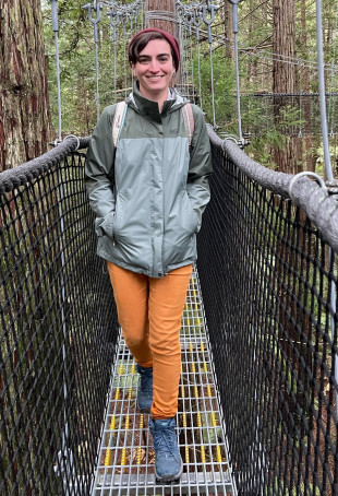 Haley posing outside on a suspension bridge, wearing a rain jacket, orange pants, and boots.