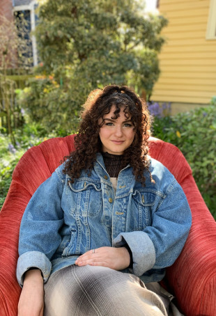 Shoshana, smiling, while reclining in a red chair in front of trees. Student-supplied profile pho...
