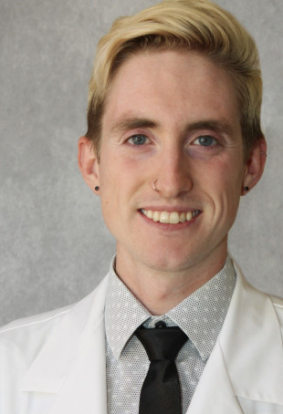 Zach smiling at the camera wearing a white medical doctor's coat, dress shirt, and black tie.