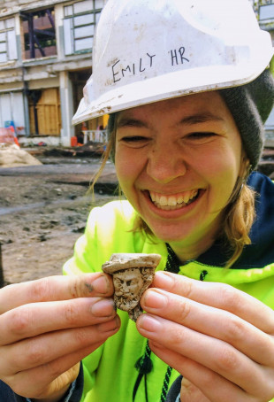 Emily, smiling while wearing construction gear, holding a 19th century pipe head found during an expedition in Australia.