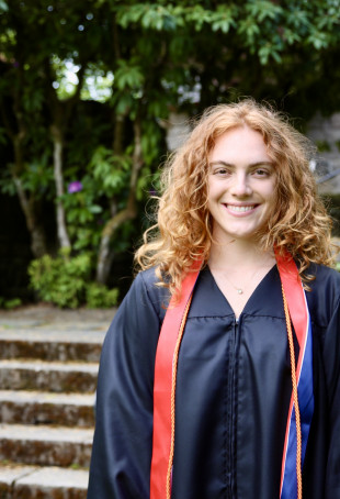 Emily Tash smiles in the campus estate gardens. She has red hair and is wearing a graduation gown.