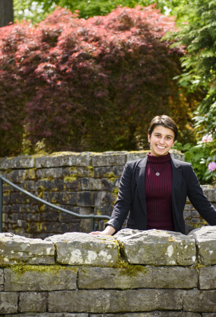 Kaylee-Anna, in a maroon turtleneck and black blazer, standing behind a stone wall in front of a ...