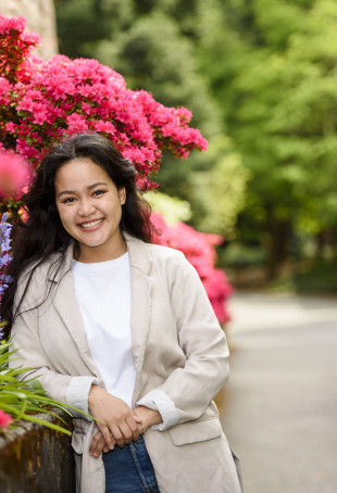 Keilani, in a beige blazer, white t-shirt and blue jeans, smiling while leaning against a stone wall next to bright, colorful flower bushes.
