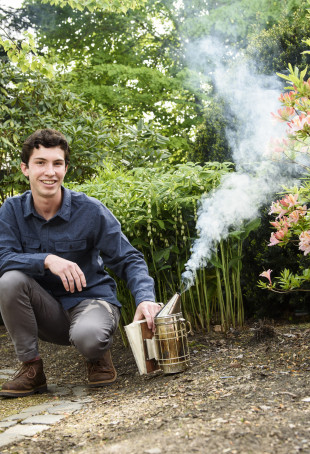 Mateo, in gray pants and a blue collared shirt, crouching on the ground in front of trees and next to a beehive smoker.