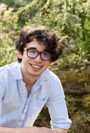 Jason, sitting on the ground in front of a garden and leaning forward in a light blue collared shirt, smiling on a sunny day.