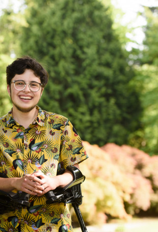 Luca, smiling while standing on a pathway, wearing a yellow collared shirt printed with a colorful bird pattern and holding arm braces.