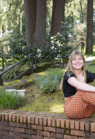 Jemma smiling outside, surrounded by trees, wearing a black top, orange patterned pants, and black boots.