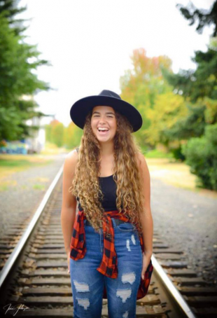 Ashtyn laughing and posing while standing on a train track, wearing a large black hat, black tank top, flannel shirt tied around waist.