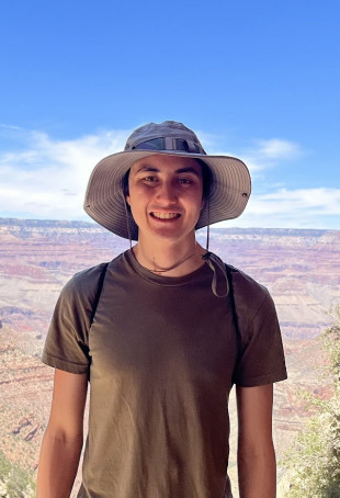 William smiling in front of the Grand Canyon, wearing a sun hat and brown t-shirt.