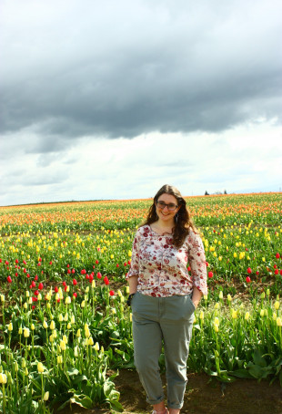 Sophie posing in front of a field of colorful wildflowers. She is wearing glasses, a shirt with flowers on it, and green pants.