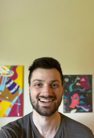 Photo of Ben in front of a wall covered in art. Ben is smiling in a gray t-shirt.