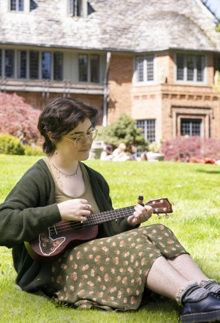 Mack sitting outside Manor House playing a ukulele. They are wearing a green floral dress with a dark green cardigan and black loafers.