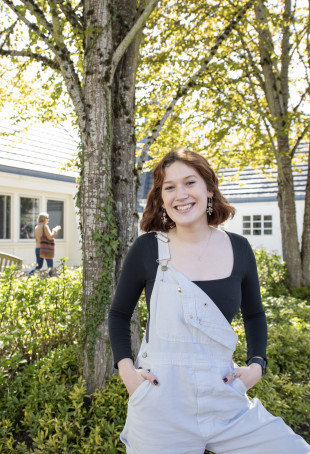 Alex posing outside. She is wearing a long-sleeved dark shirt under overalls with one strap hanging down.