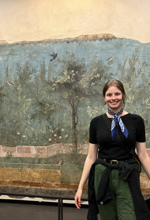 Claire Lyman posing in front of a mural wearing a black top, green pants, and a blue scarf around her neck.