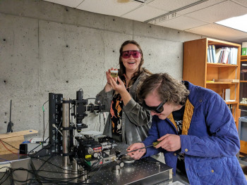 Two students wearing goggles and posing with physics research equipment.