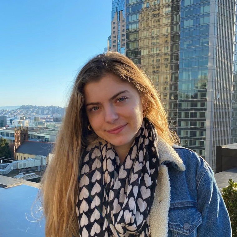 Mia, in a black and white checkered scarf and denim jacket, smiling while standing on the top of a skyscraper.