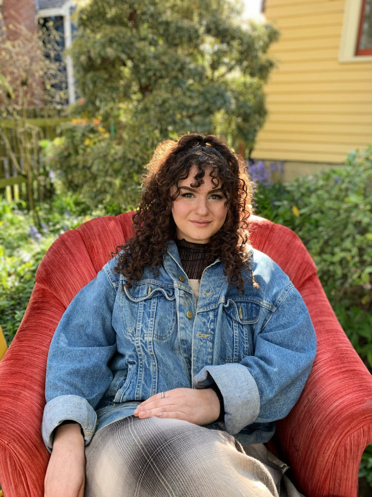 Shoshana, smiling, while reclining in a red chair in front of trees. Student-supplied profile pho...