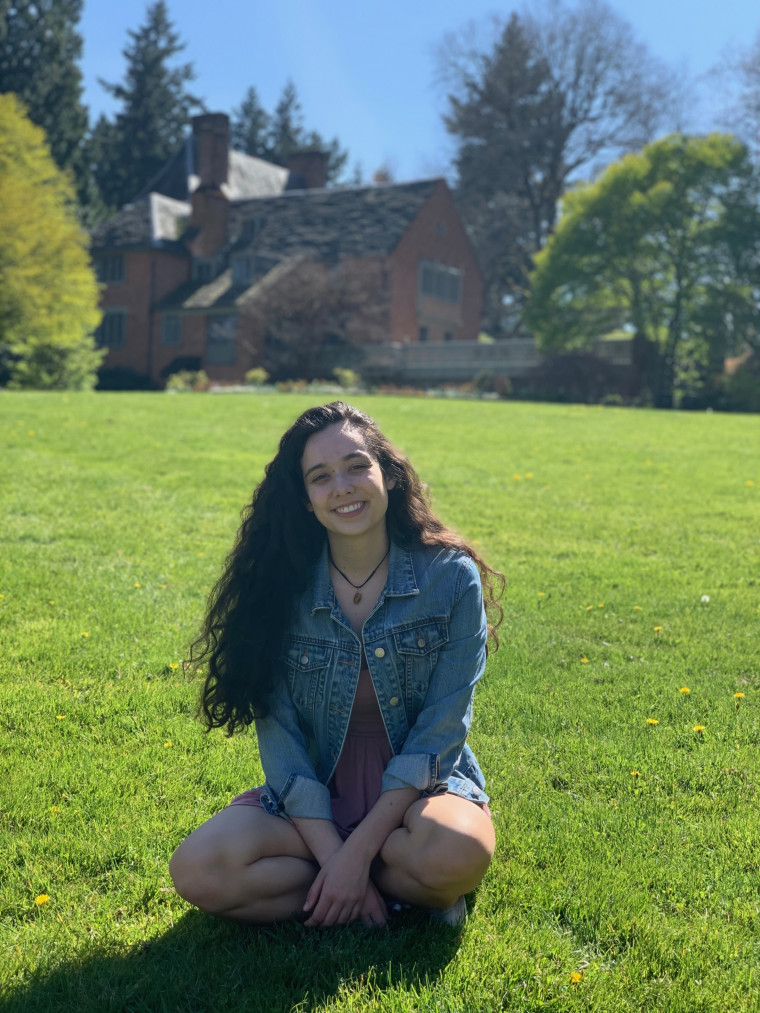 Juliana, smiling, sitting on the grass in front of the Manor House on a sunny day. Student-suppli...