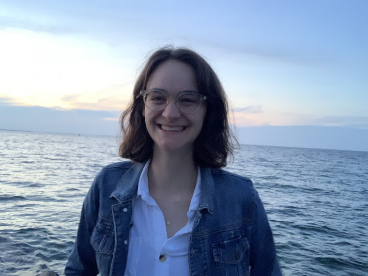 Catherine, in a white button down shirt and denim jacket, smiling while standing in front of the ocean at sunset.