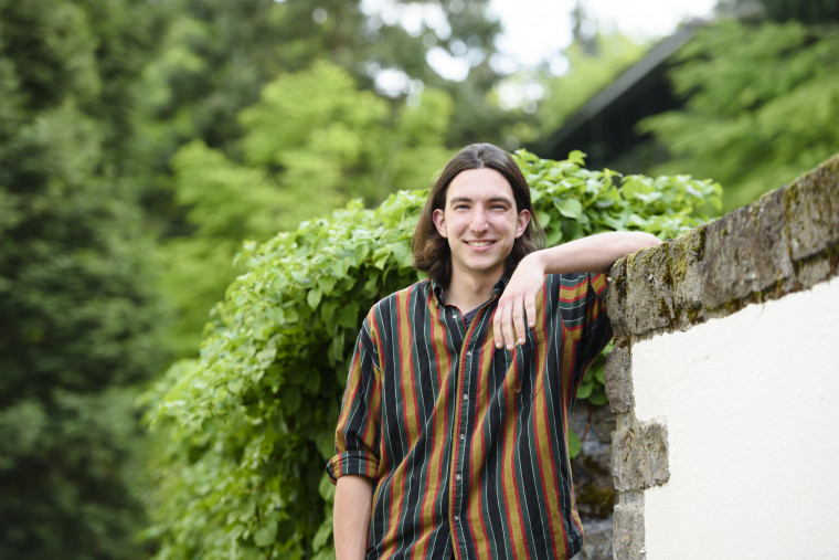 Liam, in a colorful striped collared shirt, smiling and leaning against a stone wall next to green shrubs.