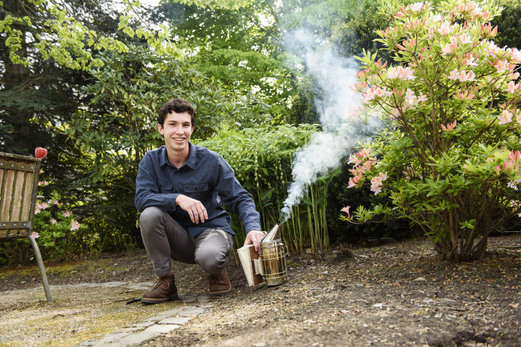 Mateo, in gray pants and a blue collared shirt, crouching on the ground in front of trees and next to a beehive smoker.