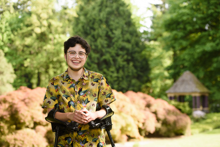 Luca, smiling while standing on a pathway, wearing a yellow collared shirt printed with a colorful bird pattern and holding arm braces.
