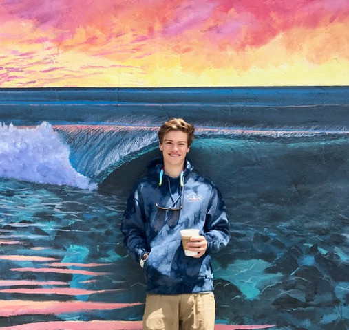 Max, holding a cup of coffee, standing in front of a wall with a painted mural of the ocean. Student-supplied profile photo due to COVID-...