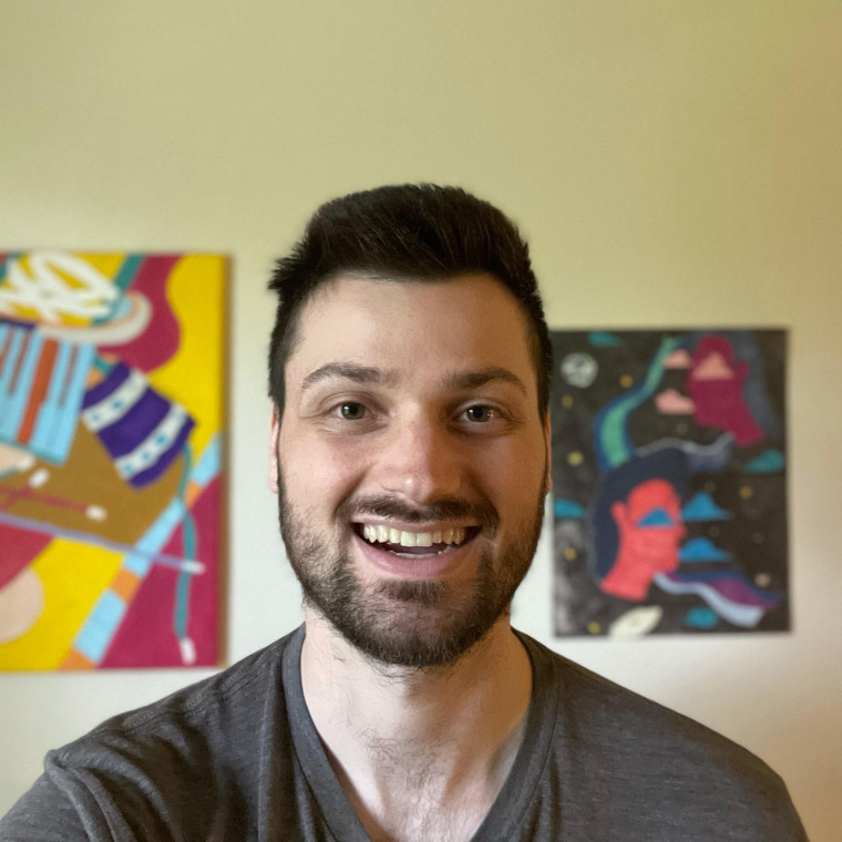 Photo of Ben in front of a wall covered in art. Ben is smiling in a gray t-shirt.