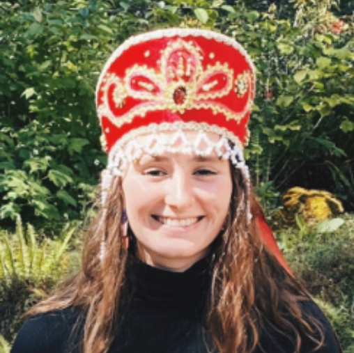 Marcella, in an elaborate red and silver Russian hat and all black outfit, smiling on a sunny day.