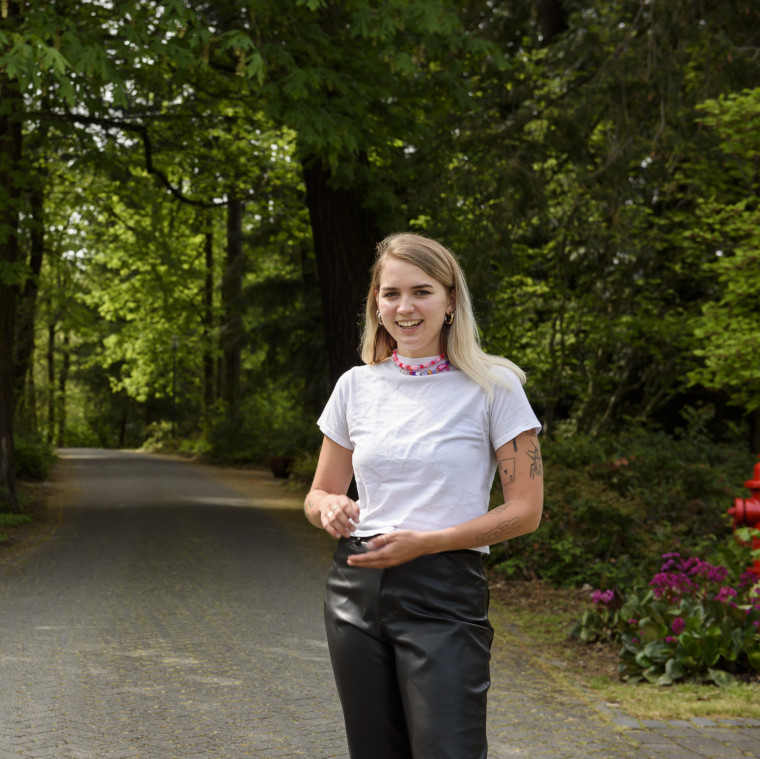 Elana, in black pants and a white t-shirt, smiling while standing in the tree-lined pathway betwe...