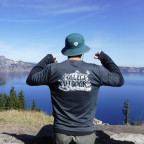 Person poses in front of Crater Lake with a teal sun hat and two thumbs pointing to the back of a grey College Outdoors shirt.