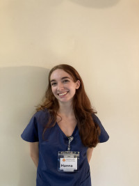 Hanna, in blue scrubs and a name tag on a lanyard, smiling and standing in front of a beige wall ...