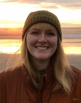 headshot of person with long blonde hair wearing a knit hat and jacket in front of a sunset