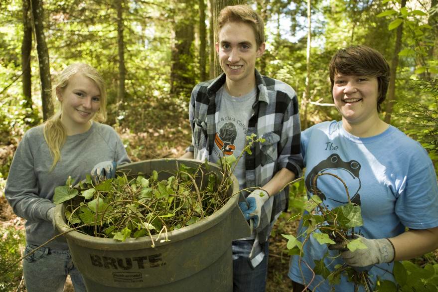 Three students standing around a refuse can and smiling while clearing weeds in the college ravin...