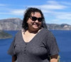 Photo of Jaime Cale - a woman standing in front of Crater Lake wearing sunglasses and a grey shir...