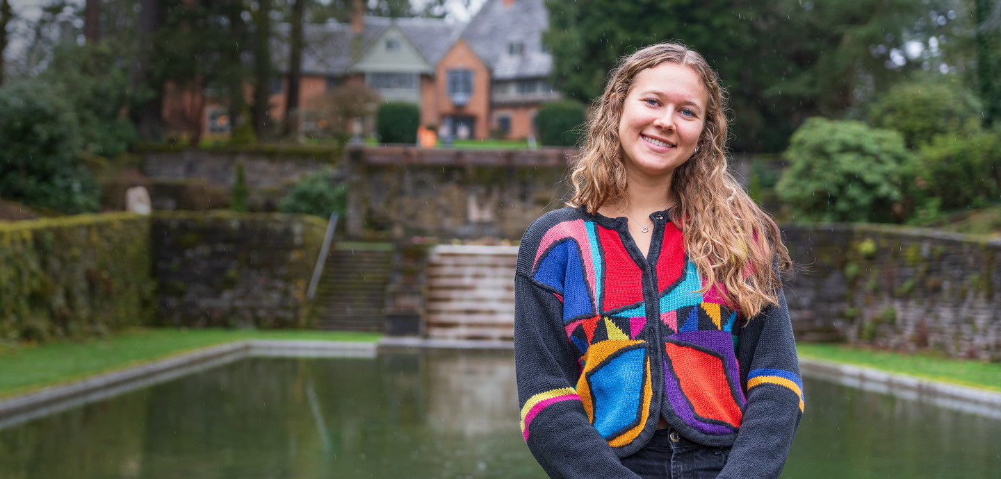 Elsa posing in front of Manor House and reflecting pool, wearing a colorful jacket.